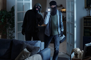 Two men wearing black ski masks entering living room at night, one man holding flashlight and bag,...