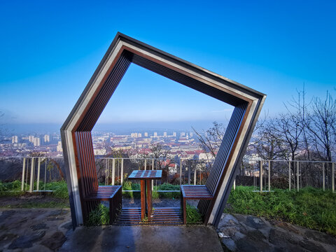 A geometric wooden bench at the Avas Lookout in Miskolc, Hungary, framing the cityscape below on a bright, clear morning.
