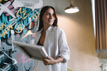 Smiling woman holds finished artwork and looks aside, embodying fulfillment, creativity and...