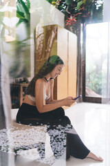 Cheerful woman with smartphone laughs while texting in modern bright home, captured through...