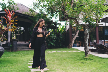 Happy young woman strolls along stone path in tropical garden, holding smartphone and...