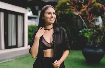 Happy woman smiling with hand near neck, dressed in black in lush tropical garden—blending...