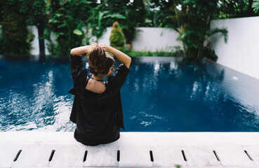 Woman adjusting hair by blue pool in black outfit, representing pause, personal care and the gentle...