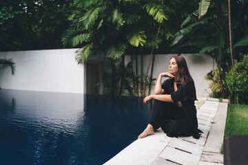 Thoughtful barefoot woman in black outfit sits at edge of infinity pool, surrounded by lush...