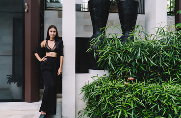 Elegant woman poses barefoot by villa, smiling subtly in tropical garden setting, merging fashion,...