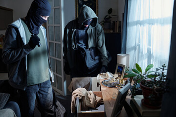 Two men wearing masks searching through drawer and examining jewelry on wooden table in residential...
