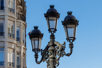 Historic street lamp standing elegantly against a clear blue sky near a building. The ornate design showcases urban architecture details in Valencia, Spain.