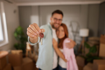 Young couple joyfully presents their new house keys while embracing each other amidst unpacked cardboard boxes in their new living space, celebrating a fresh start.