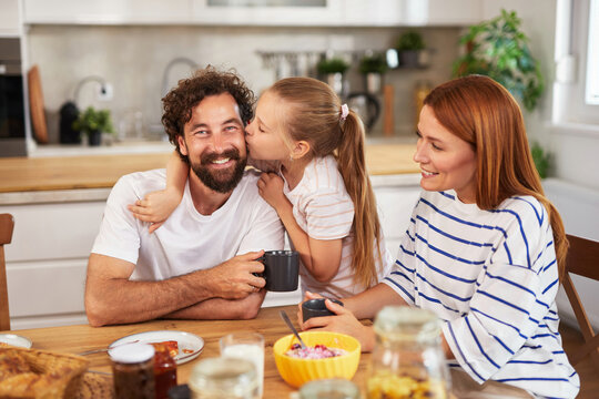A joyful family shares breakfast in a warm kitchen, with a little girl giving her dad a sweet kiss while mom smiles, creating a loving and cheerful atmosphere. - Powered by Adobe