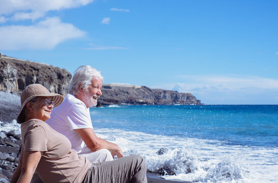 Relaxed senior couple in peaceful vacation sitting on the seashore while waves splashing on the beach enjoying retirement lifestyle and freedom, horizon over sea