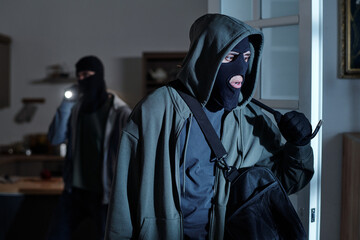 Two men wearing balaclavas entering room, one man holding bag and looking around cautiously, second...