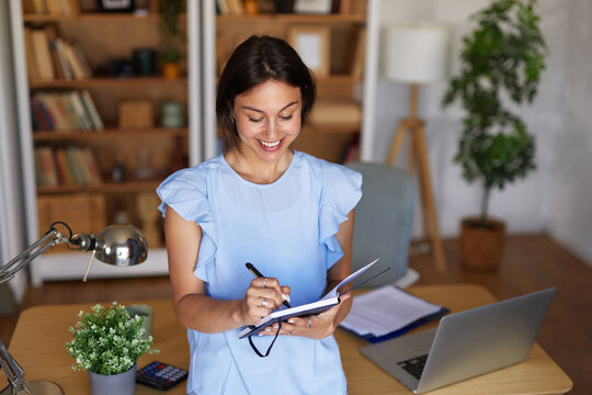 A woman stands in a contemporary home office, smiling as she writes in a notebook. A laptop and a potted plant are visible on the desk, creating a productive environment.