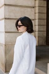 Urban woman in white blazer looking over shoulder near stone building in natural daylight