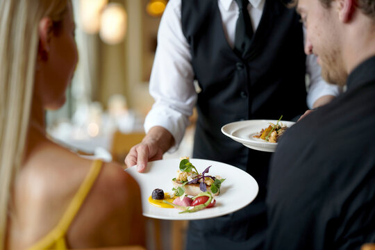Elegant waiter in black and white uniform serving a gourmet meal to a guest in a luxurious modern restaurant