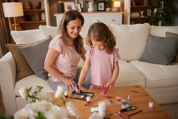 A mother and her young daughter are happily applying makeup together at a wooden table in their...