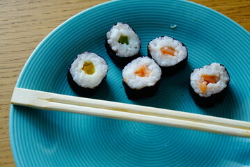 A vibrant food photograph featuring five sushi rolls arranged on a blue plate. Each roll is wrapped in seaweed and filled with white rice and colorful ingredients—yellow, green, and orange. A pair of 