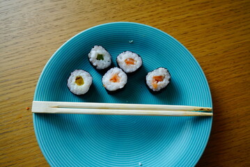A vibrant food photograph featuring five sushi rolls arranged on a blue plate. Each roll is wrapped in seaweed and filled with white rice and colorful ingredients—yellow, green, and orange. A pair of 