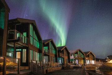 View of ethereal green and purple Northern Lights dance above cozy, warmly lit wooden cabins nestled against a dark, rocky landscape, Troms, Norway.