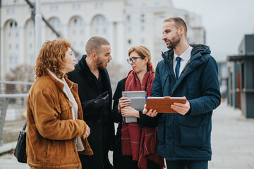 A group of four individuals conversing while standing outdoors in a city setting. They hold tablets...