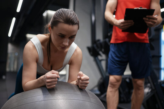 Young woman exercising with fitness ball near professional personal trainer in gym