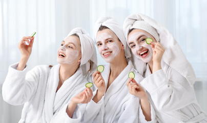 Happy women with facial masks and cucumber slices in bathrobes after spa procedures indoors