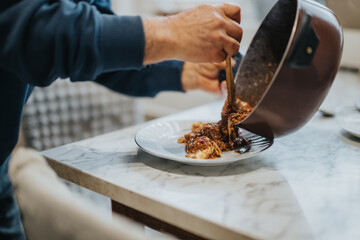 A person in a blue sweater serves a freshly prepared meal from a pot onto a plate on a marble table.