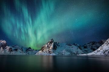 View of vibrant aurora borealis dances above snow-capped mountains, reflected in the calm waters in Norway, HamnÃ¸y, Norway.
