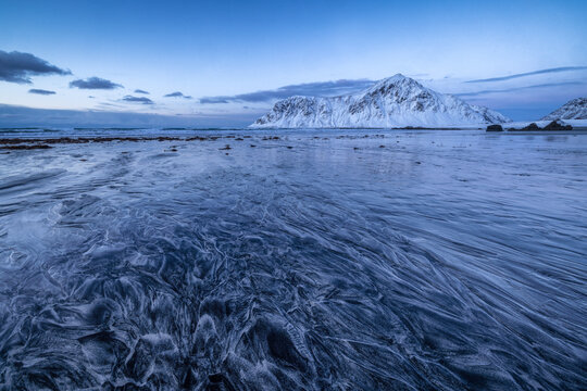 View of the patterned, wet sand reflecting the cool blue sky, contrasting with the snow-capped mountains in the distance, Unstad, Norway.