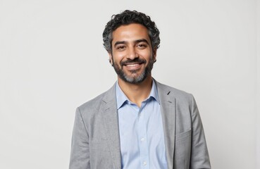 Middle-aged Saudi Arabian man with curly hair, beard smiles warmly. Wears light blue collared shirt, stylish grey blazer. Isolated on clean white background, portrait conveys confidence,
