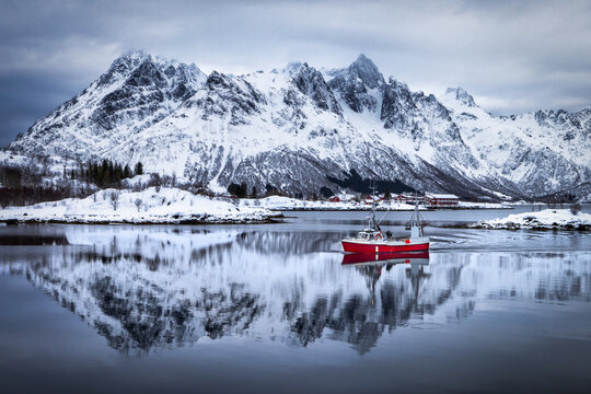 Lofoten Islands, Norway - 15 March 2023: View of a vibrant red boat cutting through the serene waters, mirroring the snow-dusted mountains under a moody sky.