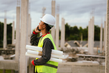 A construction worker wearing a yellow helmet and safety vest examines blueprints on a building...