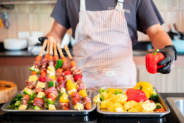 Chef holding a red bell pepper while preparing colorful meat and vegetable skewers, showcasing fresh ingredients and vibrant food preparation in a home kitchen setting.