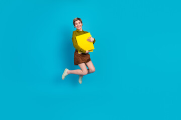 Fototapeta premium Confident young professional woman holding yellow folders while jumping against a vibrant blue background.