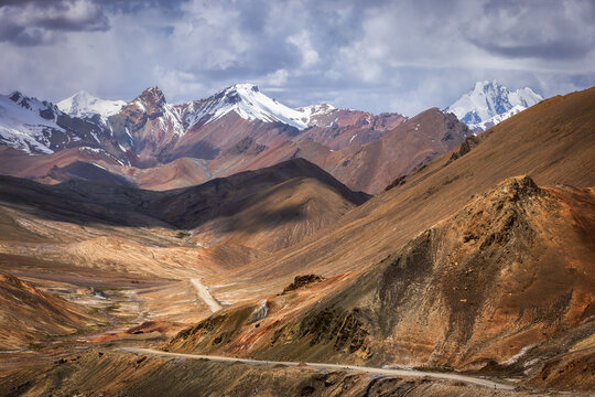 View of winding road cutting through rugged, sun-drenched valleys, contrasting with the snow-capped peaks under a dramatic sky, ak baital, Tajikistan.