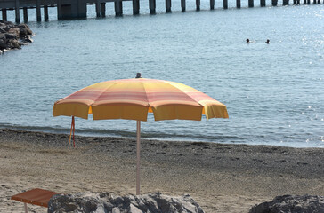 Ombrellone giallo-arancio e lettino vuoto su una spiaggia di ciottoli del Mediterraneo con sfondo del molo.