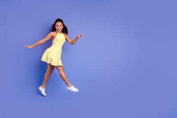 Fototapeta premium Young cheerful woman in a yellow dress smiling energetically, posing mid-jump against a vibrant violet background