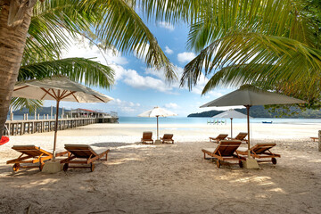 Wooden lounge chairs under beach umbrellas on a pristine white sand beach, offering a tranquil escape near a tropical pier under a sunny sky.
