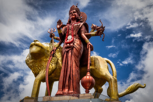 View of a copper-colored Durga statue stands tall upon a golden lion amidst a vibrant sky, holding weapons with multiple arms, Ganga Talao, Mauritius.