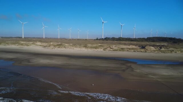 Drone slide left over a sandy beach with wind farm nin the background on a sunny day.