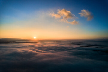 Aerial view of sun rises above dense clouds, casting warm, golden glow across horizon. Scene captures tranquility of new day, as the first rays of sunlight break through, creating serene atmosphere.