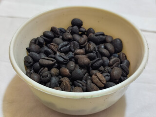 A bowl of freshly roasted coffee beans is dark in color, on a white background.