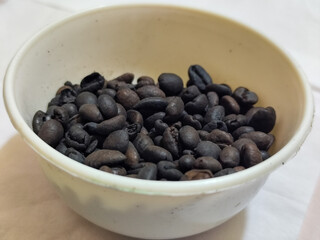 A bowl of freshly roasted coffee beans is dark in color, on a white background.
