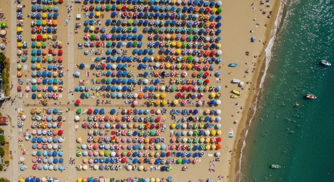 Aerial view shows a crowded beach, vibrant rows of parasols providing shade for sunbathers next to turquoise water. Small boats float nearby - Powered by Adobe