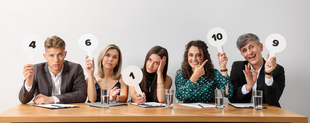 Panel of emotional judges voting with score signs at table against light grey background
