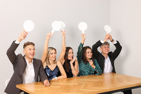 Panel of happy judges voting with blank score signs at table near grey wall