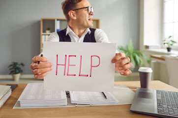 Upset overworked young man accountant sitting at the desk at his workplace in office with a pile of...