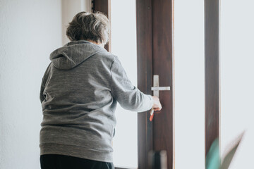 An elderly woman wearing a grey hoodie opens a wooden door, allowing bright natural light to enter...