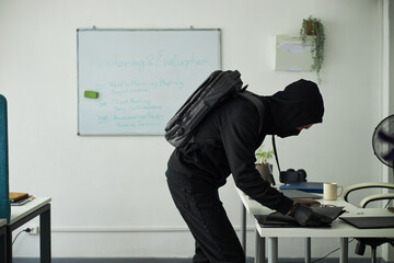 Young man wearing black outfit, face mask, backpack and gloves bending over corporate office desk searching through documents in modern workspace and stealing laptop