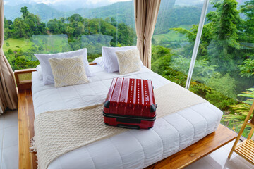 Red suitcase placed on a cozy white bed in a resort room with panoramic forest and mountain view outside glass windows. Perfect start to a relaxing nature getaway.