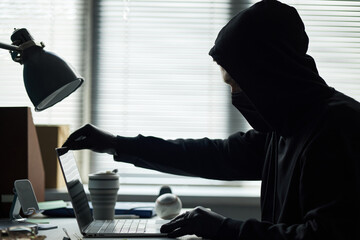 Young male intruder wearing black hoodie, face mask, gloves sitting at desk using laptop, breaking...